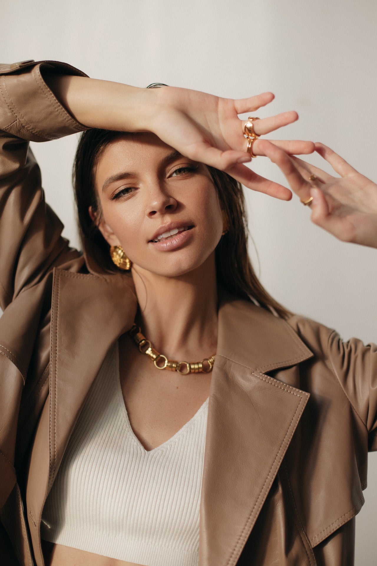 Stylish female model wearing a light brown leather jacket, white crop top and golden necklace, earrings, and rings.